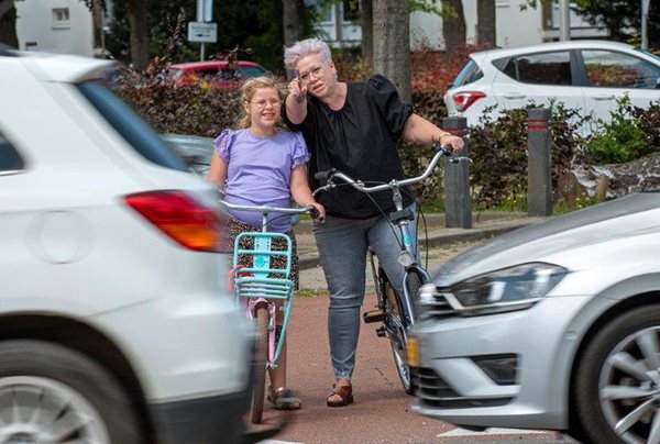 Wies met een paars shirt op haar fiets samen me teen vrouw. Ze staan achter twee grijze auto's.