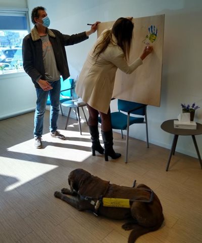 Laura zet handtekening op schilderij. Hond ligt achter haar. Man met mondkap staat naast haar.