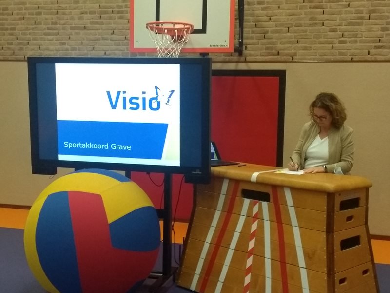 Vrouw met blond krullend haar staat in de gymzaal naast heet scherm met de presentatie waar sportakkoord Grave staat beschreven.