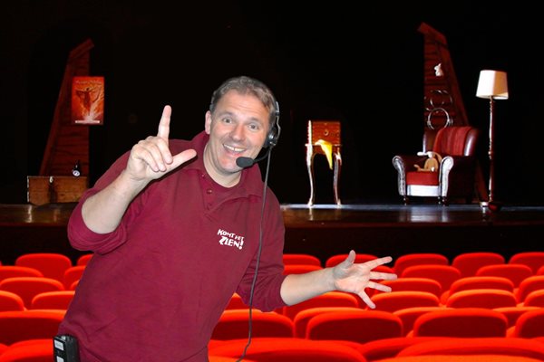 Hilbert op de foto met zijn handen omhoog. 1 hand in een l-vorm en de andere hand wijd open. Hij staat in de theaterzaal die rood is van kleur.