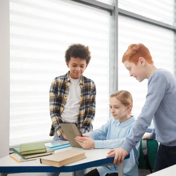 Drie leerlingen werken samen aan een schootafeltje, een meisje zit met een iPad, twee jongens staan ernaast Drie leerlingen werken samen aan een schootafeltje, een meisje zit met een iPad, twee jongens staan ernaast