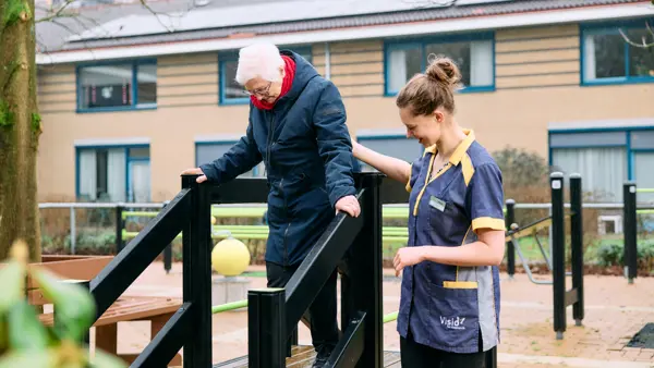 Cliënt loopt buiten voorzichtig over een smalle oefenbrug met leuningen, terwijl een begeleider van Visio De Vlasborch naast de cliënt meeloopt Cliënt loopt buiten voorzichtig over een smalle oefenbrug met leuningen, terwijl een begeleider van Visio De Vlasborch naast de cliënt meeloopt