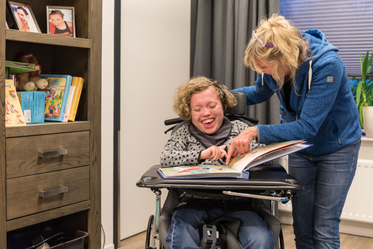 Medewerker leest een groot boek samen met een cliënt in een rolstoel, waarbij ze dingen aanwijzen in het boek