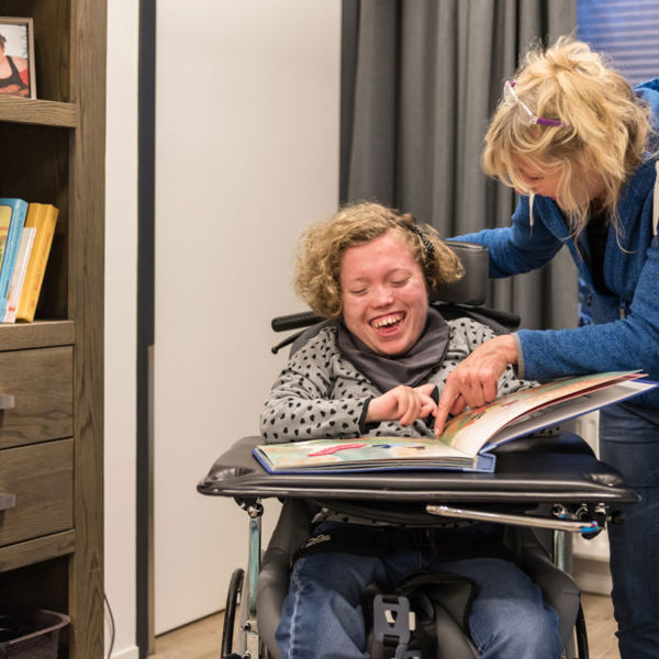 Medewerker leest een groot boek samen met een cliënt in een rolstoel, waarbij ze dingen aanwijzen in het boek Medewerker leest een groot boek samen met een cliënt in een rolstoel, waarbij ze dingen aanwijzen in het boek