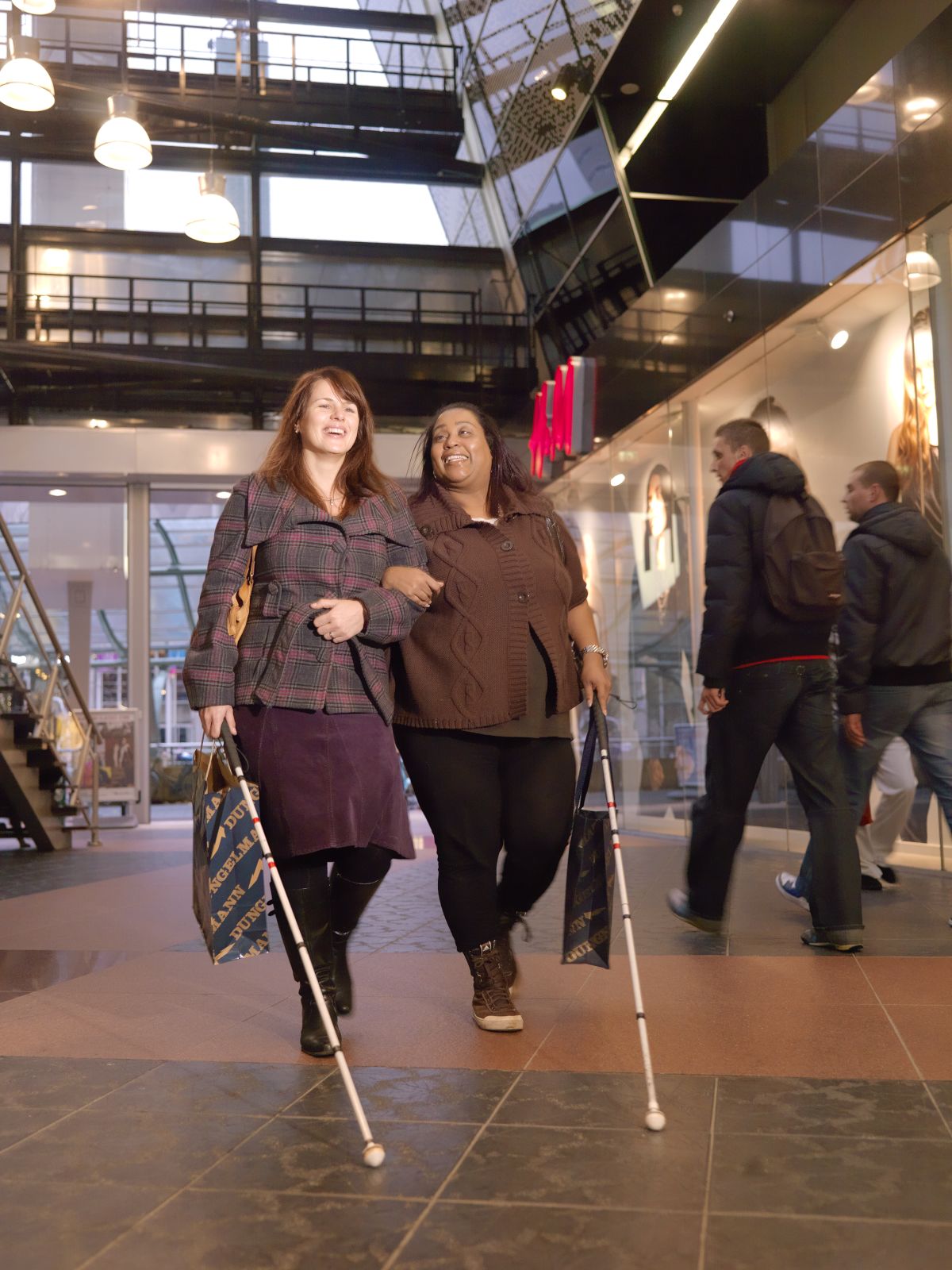 Twee vrouwen lopen door een gebouw. Ze hebben allebei een taststok vast. 