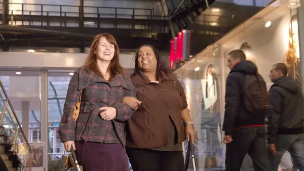 Twee vrouwen lopen door een gebouw. Ze hebben allebei een taststok vast. Twee vrouwen lopen door een gebouw. Ze hebben allebei een taststok vast.