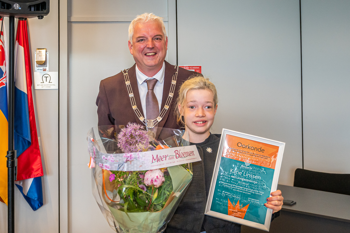 Burgemeester staat naast een kind dat een oorkonde en een grote bos bloemen vasthoudt in een binnenruimte.