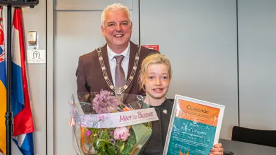 Burgemeester staat naast een kind dat een oorkonde en een grote bos bloemen vasthoudt in een binnenruimte. Burgemeester staat naast een kind dat een oorkonde en een grote bos bloemen vasthoudt in een binnenruimte.