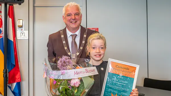 Burgemeester staat naast een kind dat een oorkonde en een grote bos bloemen vasthoudt in een binnenruimte. Burgemeester staat naast een kind dat een oorkonde en een grote bos bloemen vasthoudt in een binnenruimte.