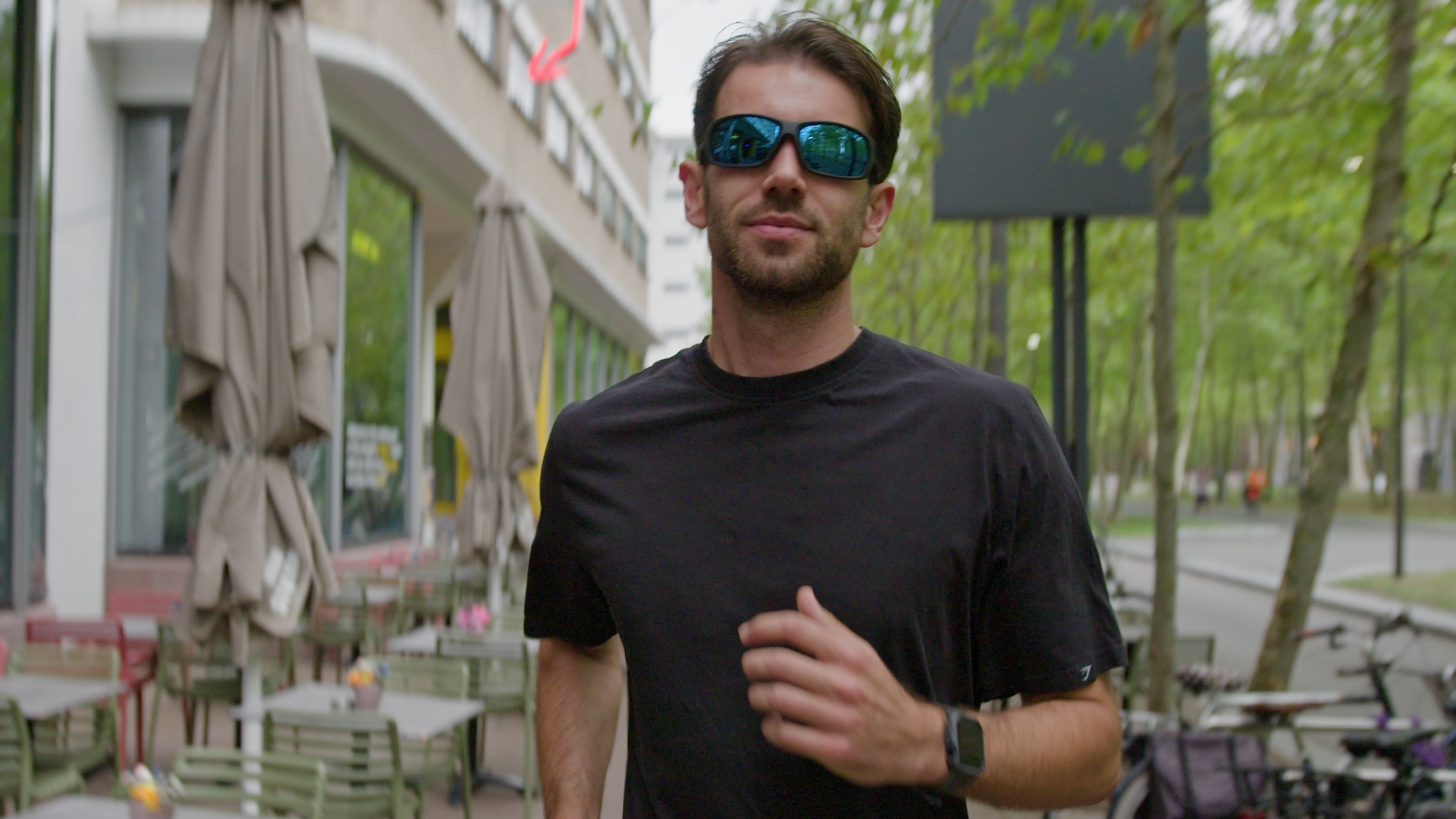 Joost is aan het hardlopen. Hij draagt een zwart sportshirt en een zonnebril. Hij rent richting te camera. 