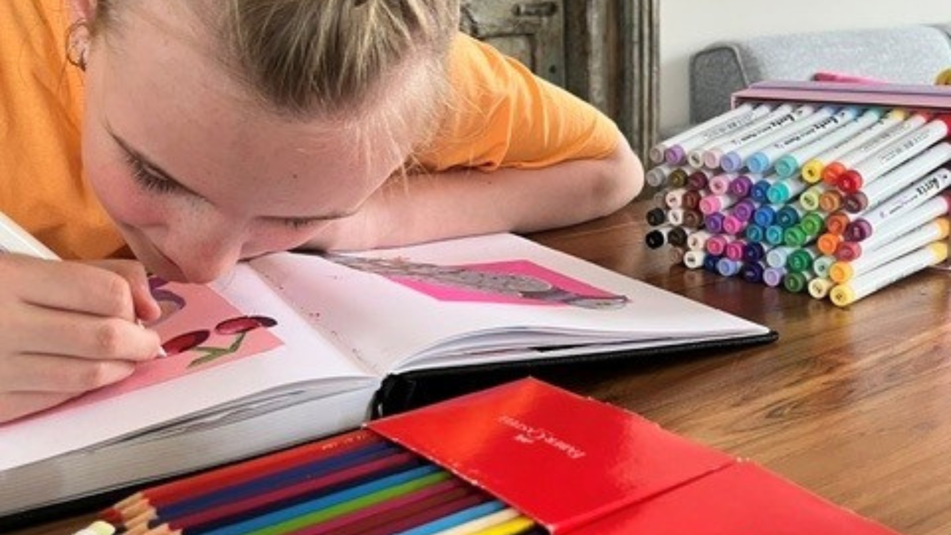 Liv zit aan een tafel en maakt een tekening in een schetsboek, omringd door stiften en kleurpotloden.