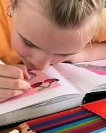 Liv zit aan een tafel en maakt een tekening in een schetsboek, omringd door stiften en kleurpotloden. Liv zit aan een tafel en maakt een tekening in een schetsboek, omringd door stiften en kleurpotloden.