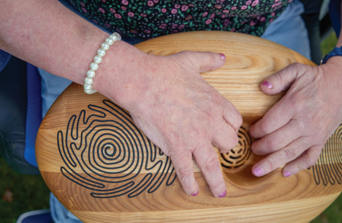 Twee handen houden de CRDL vast. Het is een houten ovaal met gegrafeerde lijnen erop. 