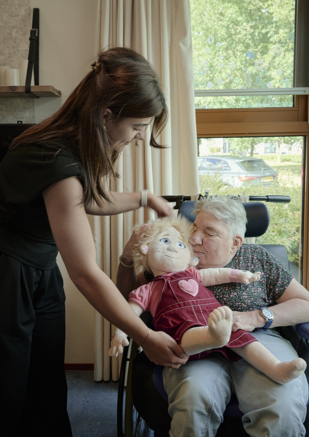 Cliënt zit in een rolstoel en houdt een grote pop op schoot, terwijl een begeleider helpt de pop goed vast te houden in een lichte kamer bij het raam. 