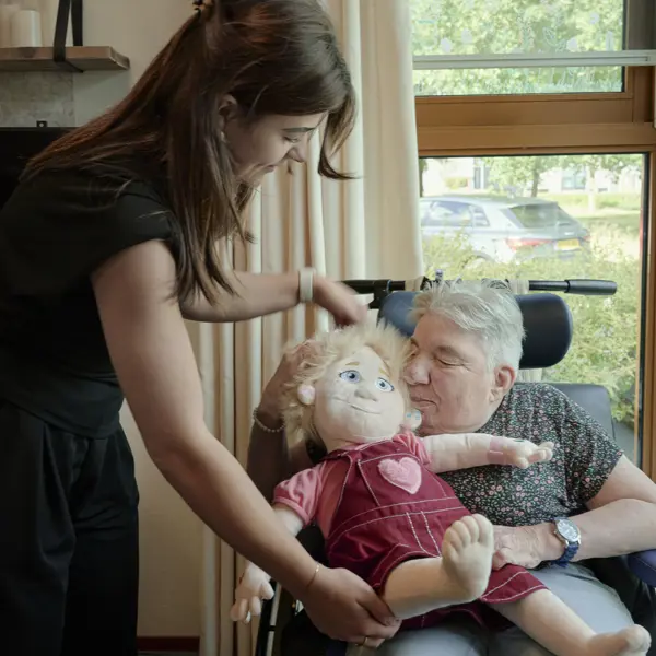 Cliënt zit in een rolstoel en houdt een grote pop op schoot, terwijl een begeleider helpt de pop goed vast te houden in een lichte kamer bij het raam. Cliënt zit in een rolstoel en houdt een grote pop op schoot, terwijl een begeleider helpt de pop goed vast te houden in een lichte kamer bij het raam.