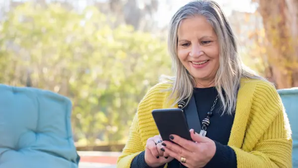 Oudere dame zit buiten in de zon en bedient haar smartphone, ze glimlacht Oudere dame zit buiten in de zon en bedient haar smartphone, ze glimlacht