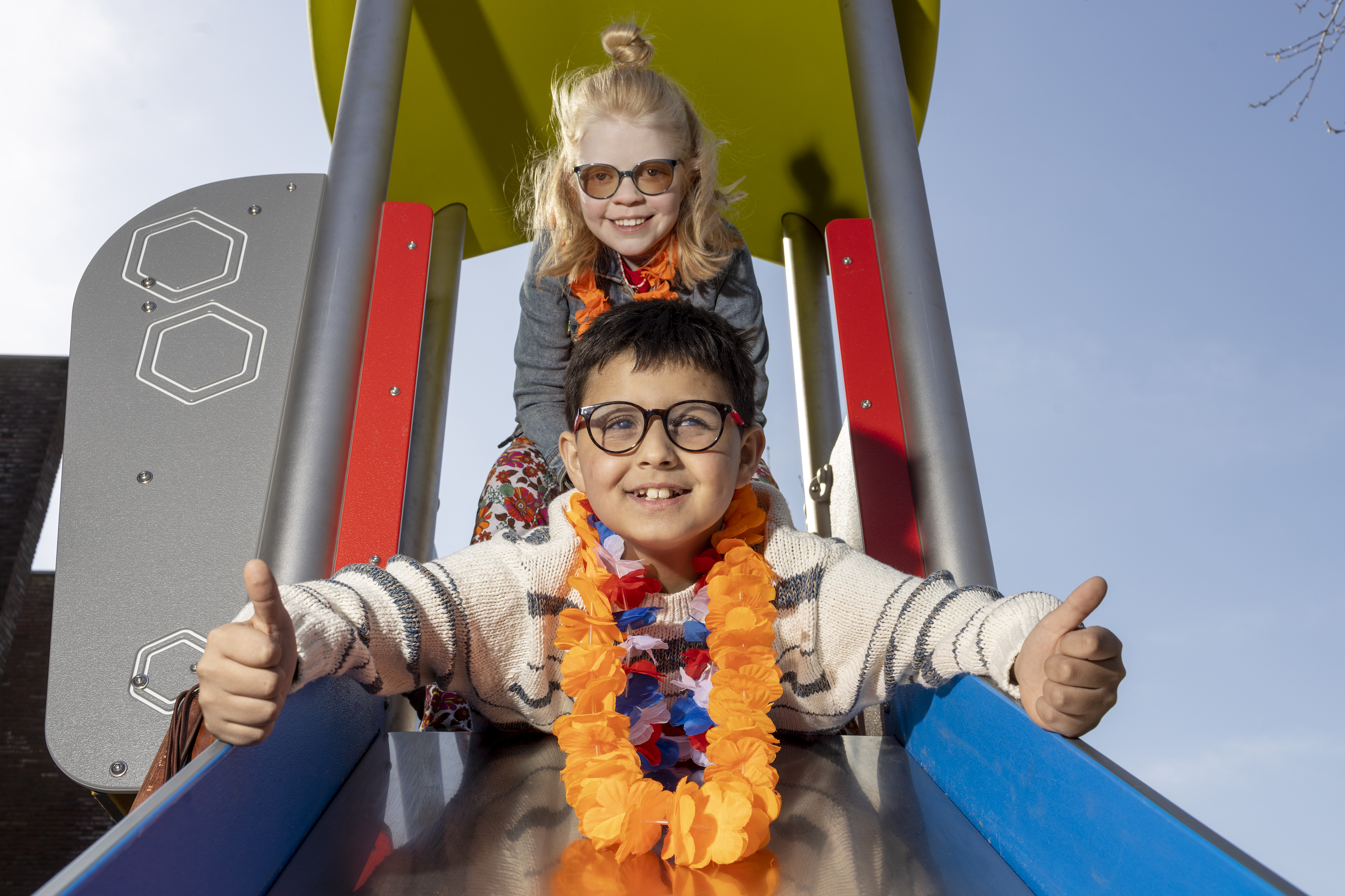 Twee kinderen, een jongen en een meisje, gaan samen van de glijbaan af. De jongen steekt zijn duimen omhoog. 