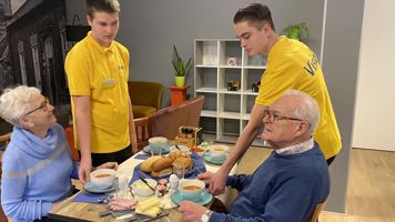 Twee leerlingen bedienen twee oudere klanten die lunchen bij huiskamercafé Visit Twee leerlingen bedienen twee oudere klanten die lunchen bij huiskamercafé Visit