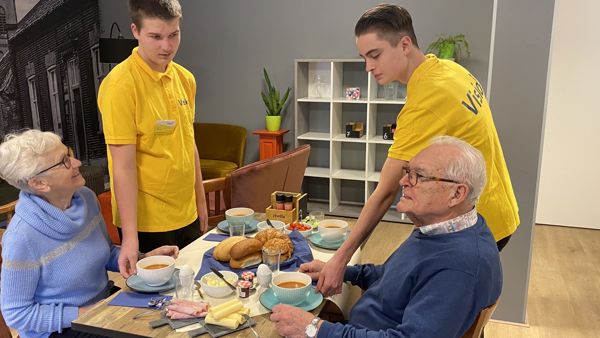 Twee leerlingen bedienen twee oudere klanten die lunchen bij huiskamercafé Visit Twee leerlingen bedienen twee oudere klanten die lunchen bij huiskamercafé Visit