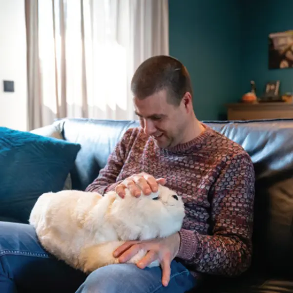Cliënt zit op de bank met Paro de zeehond op zijn schoot. Cliënt zit op de bank met Paro de zeehond op zijn schoot.