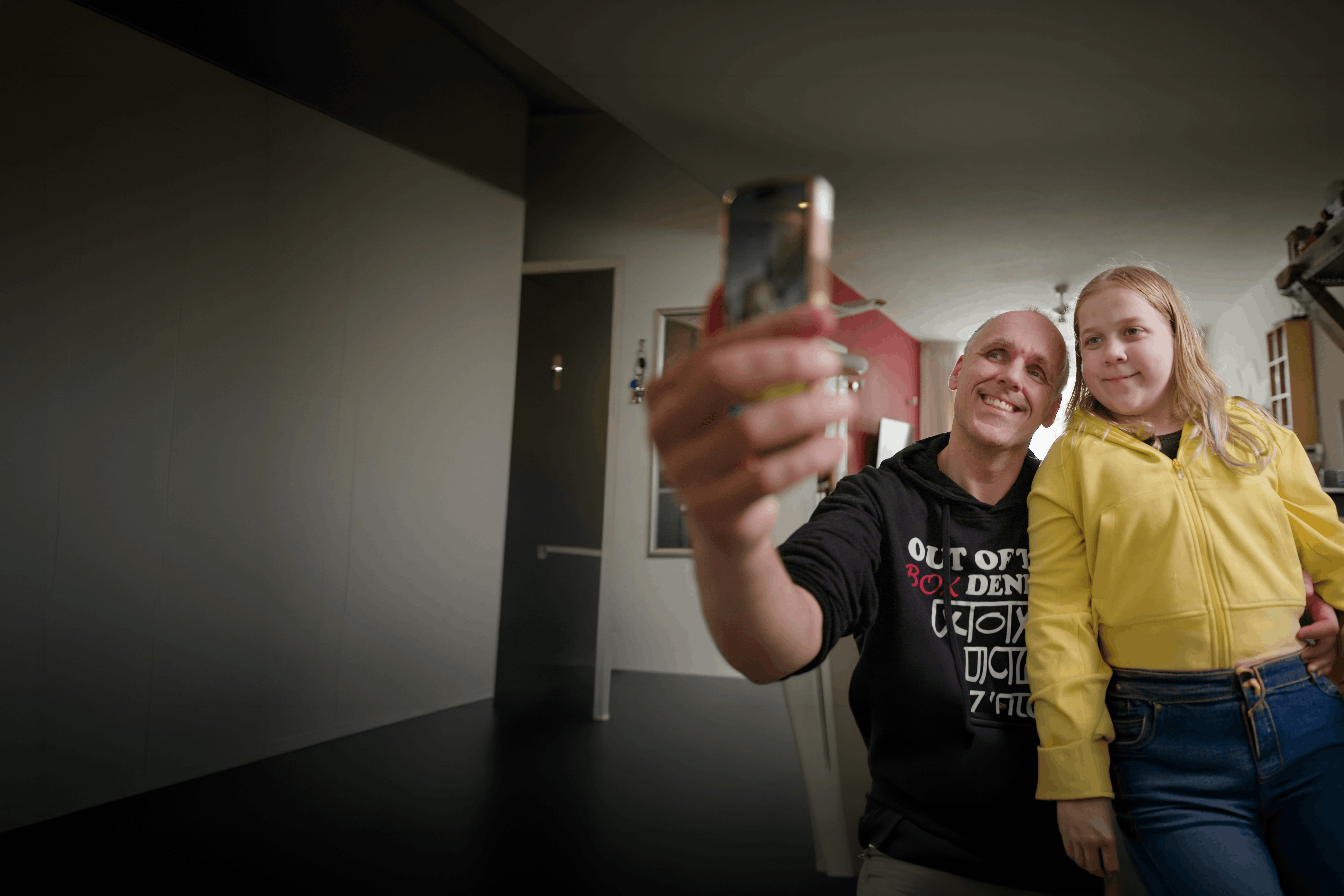 Een slechtziende man van middelbare leeftijd en zijn dochter van ongeveer 13 jaar maken samen een selfie. Ze kijken allebei lachend naar de camera van de telefoon. 