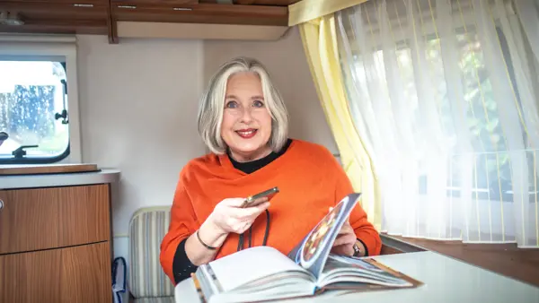 Tineke zit in een caravan aan tafel. Ze maakt een foto van een kookboek met haar telefoon. Tineke zit in een caravan aan tafel. Ze maakt een foto van een kookboek met haar telefoon.