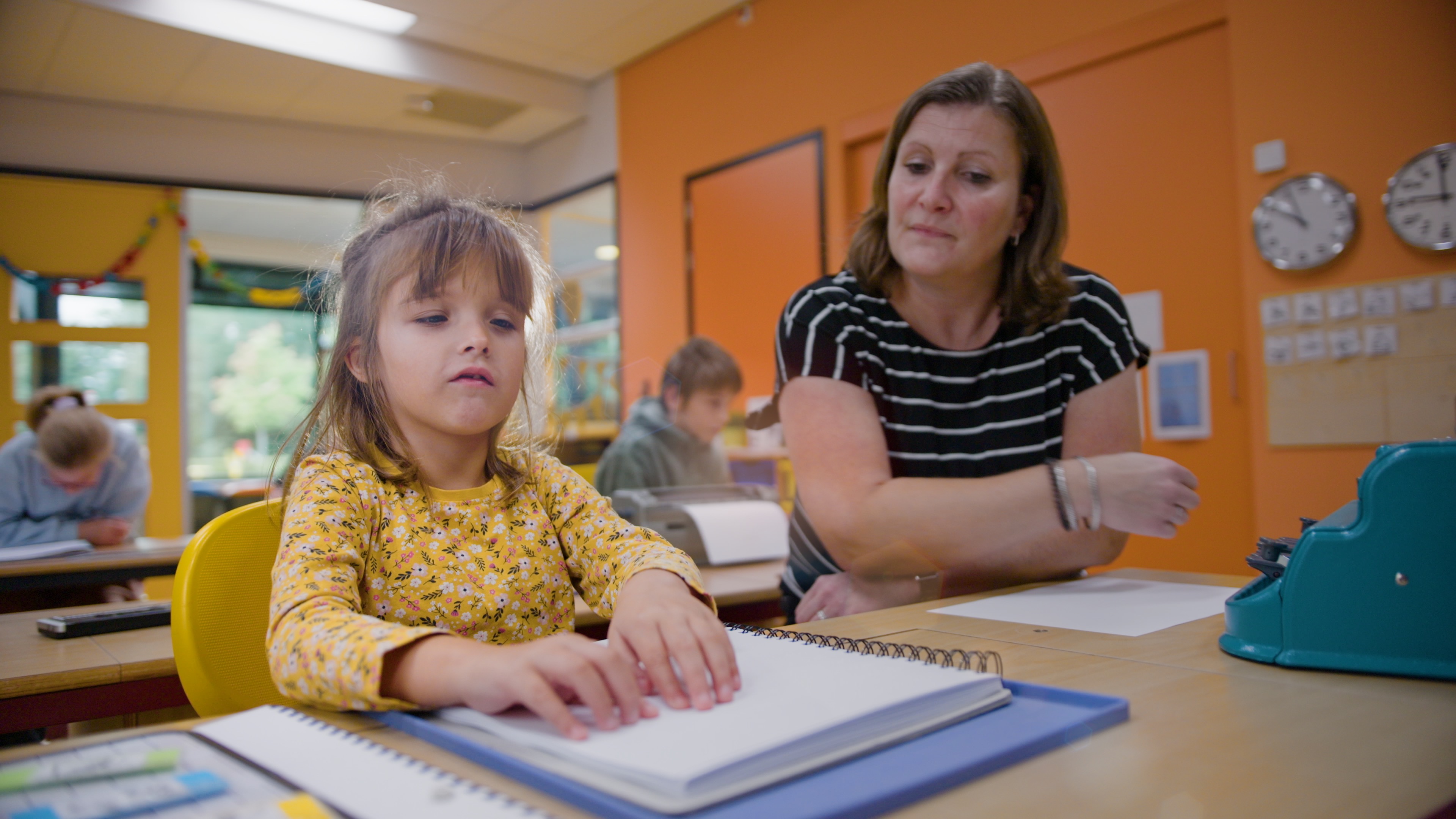 Elena zit in de klas naast de juf. Ze is braille aan het lezen. 