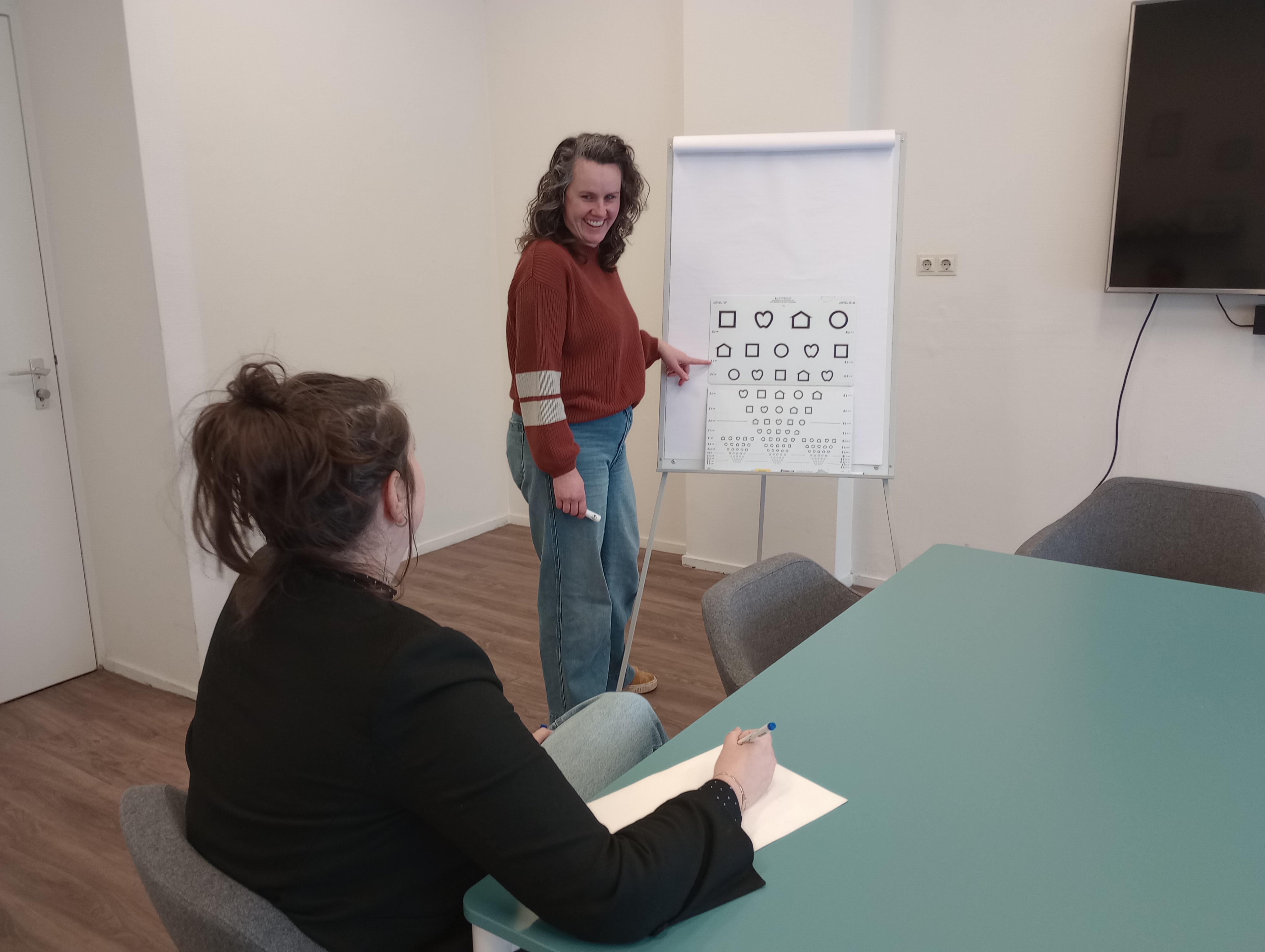 Medewerker van Visio staat voor een bord en wijst naar de symbolen die daar op staan. Ze legt iets uit aan een andere persoon, die naar het bord kijkt. 