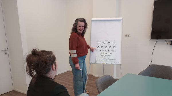 Medewerker van Visio staat voor een bord en wijst naar de symbolen die daar op staan. Ze legt iets uit aan een andere persoon, die naar het bord kijkt. Medewerker van Visio staat voor een bord en wijst naar de symbolen die daar op staan. Ze legt iets uit aan een andere persoon, die naar het bord kijkt.