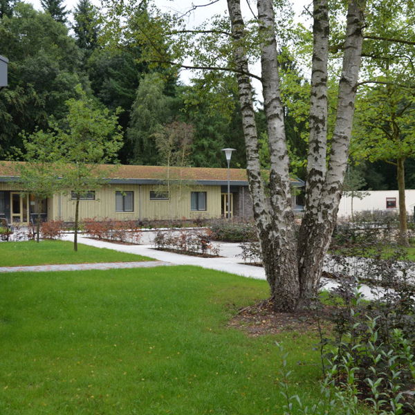 Sfeerimpressie van het groene terrein van De Blauwe Kamer met op de achtergrond de woningen en op de voorgrond veel bomen en gras Sfeerimpressie van het groene terrein van De Blauwe Kamer met op de achtergrond de woningen en op de voorgrond veel bomen en gras