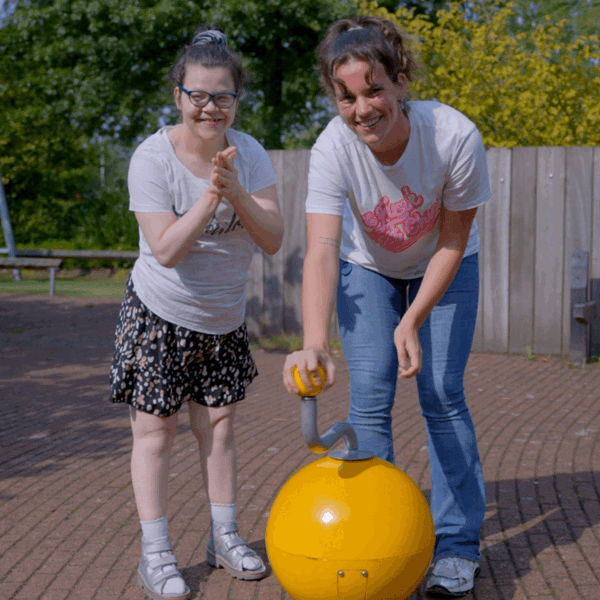 Cliënt met visuele en verstandelijke beperking en begeleider poseren bij een speeltoestel Cliënt met visuele en verstandelijke beperking en begeleider poseren bij een speeltoestel