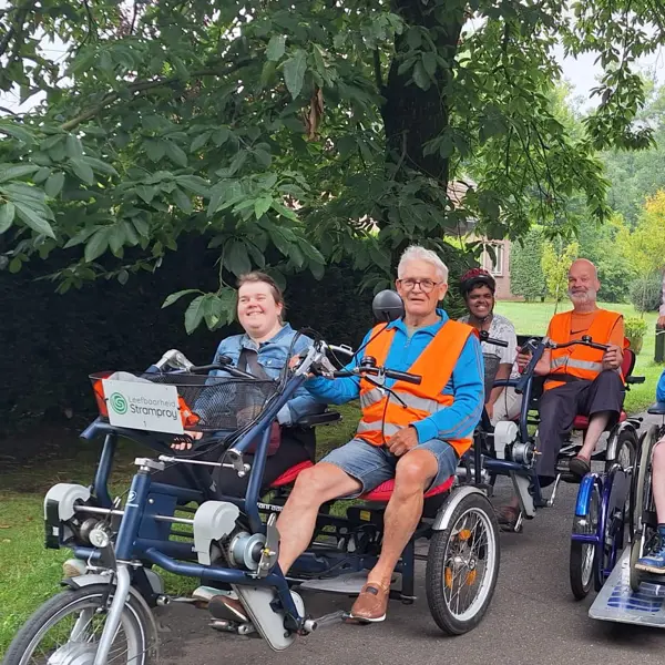 Een aantal vrijwilligers fietsen samen met cliënten in tweepersoonsfietsen. Een aantal vrijwilligers fietsen samen met cliënten in tweepersoonsfietsen.