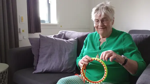 Foto van Sannie die met een breiring creatief aan de slag is, zij draagt een broek met een groen-wit patroon, een groene blouse met mouwen en een bril. Foto van Sannie die met een breiring creatief aan de slag is, zij draagt een broek met een groen-wit patroon, een groene blouse met mouwen en een bril.