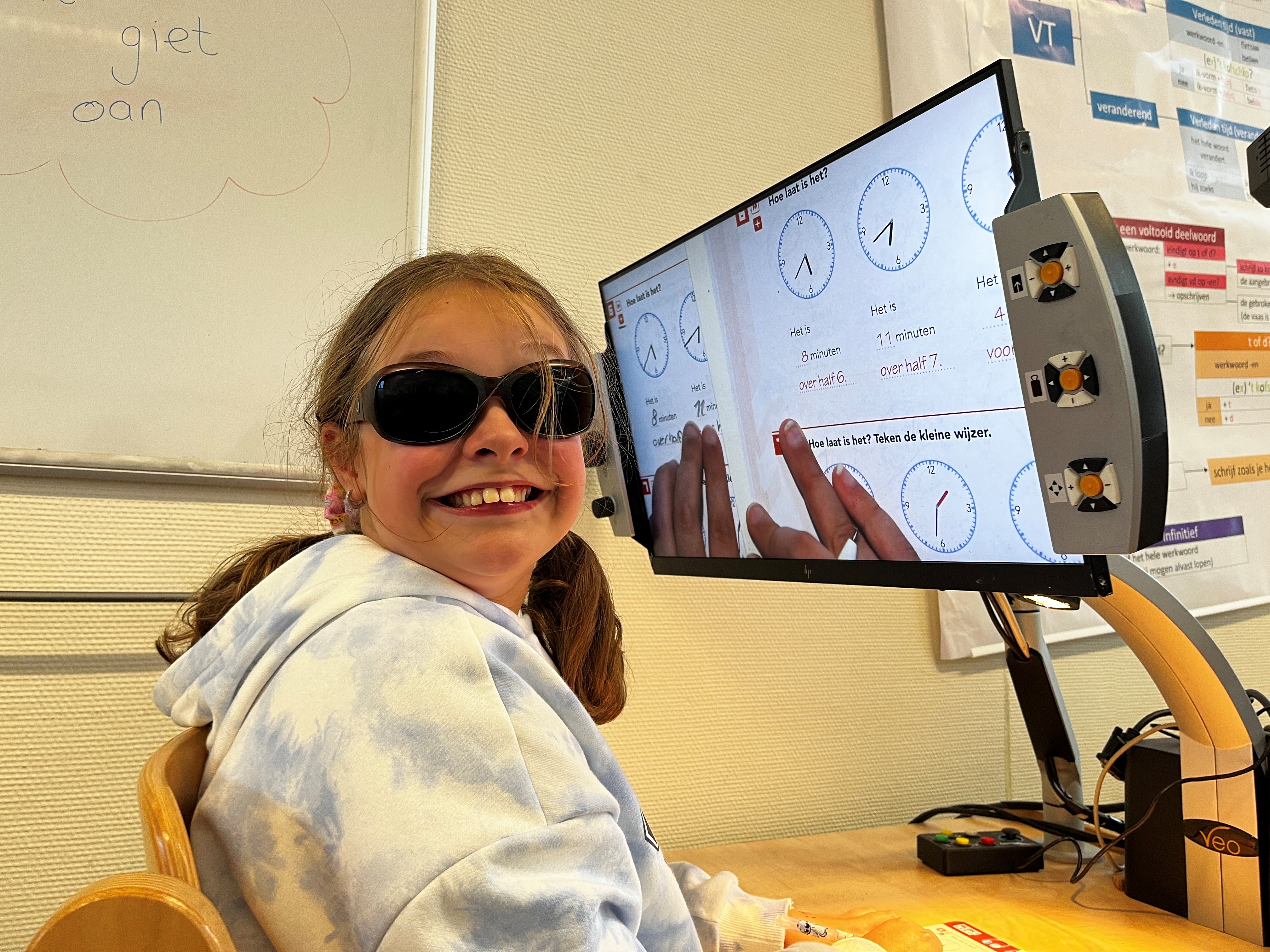 Laura glimlacht naar de camera. Ze draag een zonnebril. Ze zit voor een beeldscherm waarop haar uitvergrootte werkboek te zien is. 
