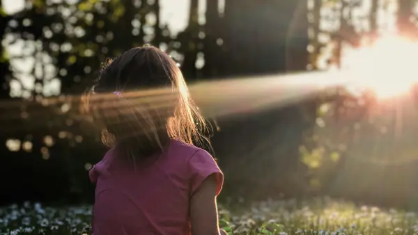 Een meisje zit in het bos op het gras. De zon schijnt door de bomen heen, ze kijkt richting het licht. Een meisje zit in het bos op het gras. De zon schijnt door de bomen heen, ze kijkt richting het licht.