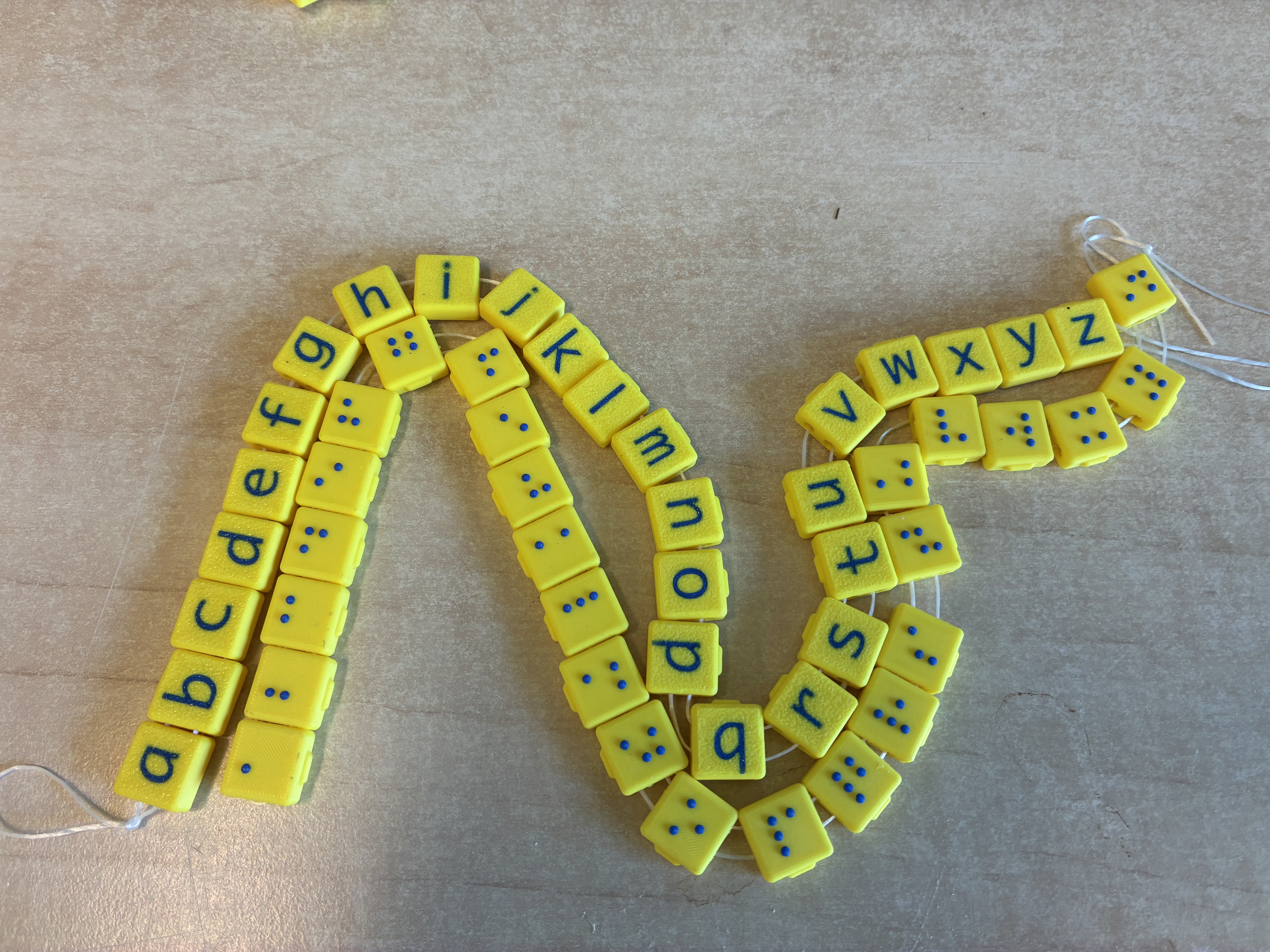 Twee koorden met gele, 3D-geprinte blokjes: één met letters van het alfabet en één met de bijbehorende brailletekens, als tastbaar leermiddel voor kinderen met een visuele beperking.