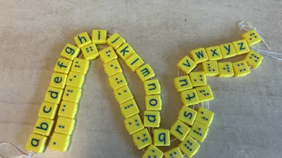 Twee koorden met gele, 3D-geprinte blokjes: één met letters van het alfabet en één met de bijbehorende brailletekens, als tastbaar leermiddel voor kinderen met een visuele beperking. Twee koorden met gele, 3D-geprinte blokjes: één met letters van het alfabet en één met de bijbehorende brailletekens, als tastbaar leermiddel voor kinderen met een visuele beperking.
