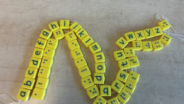 Twee koorden met gele, 3D-geprinte blokjes: één met letters van het alfabet en één met de bijbehorende brailletekens, als tastbaar leermiddel voor kinderen met een visuele beperking. Twee koorden met gele, 3D-geprinte blokjes: één met letters van het alfabet en één met de bijbehorende brailletekens, als tastbaar leermiddel voor kinderen met een visuele beperking.