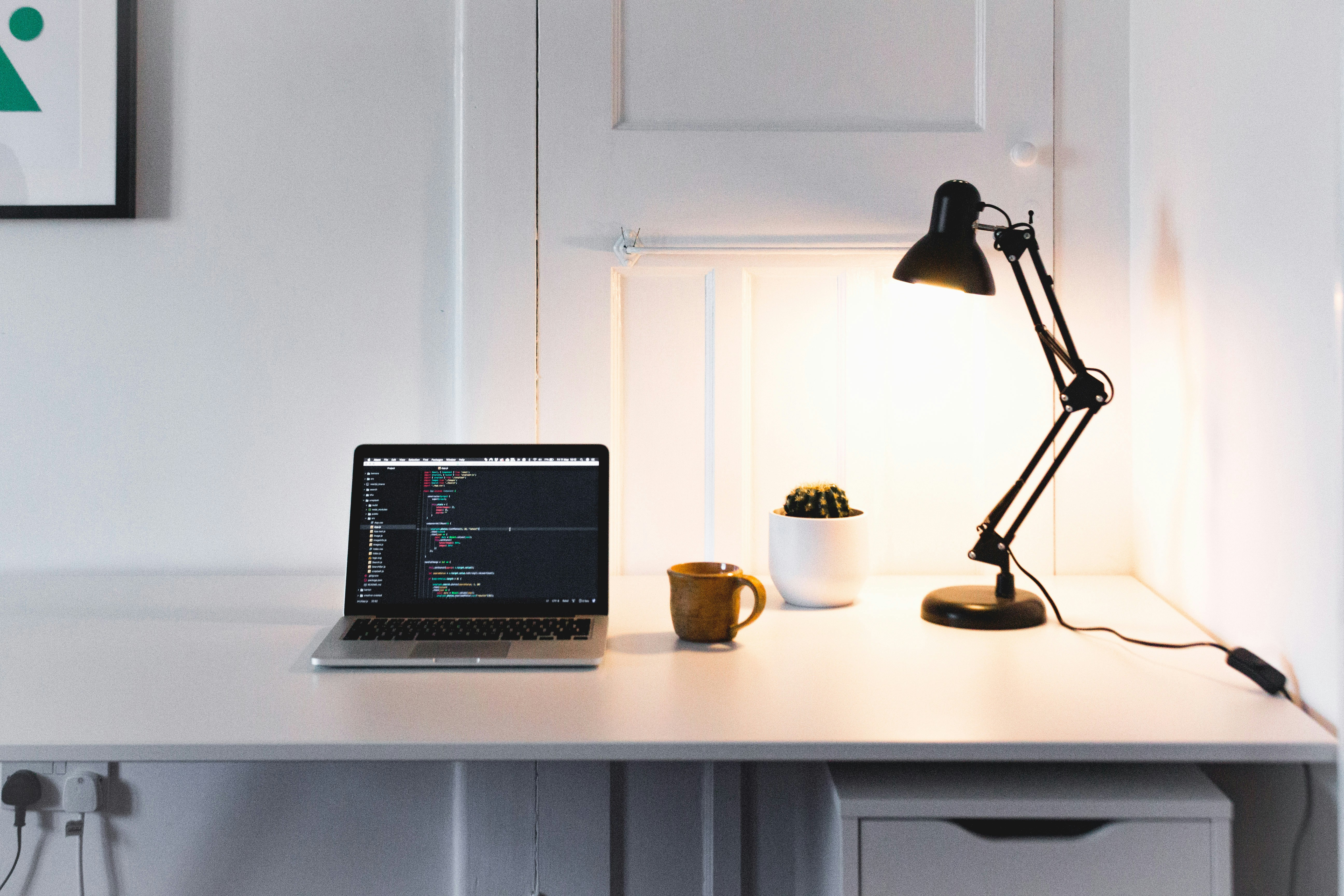 Minimalistische werkplek met een laptop op een wit bureau, naast een mok, een kleine cactus en een brandende bureaulamp.