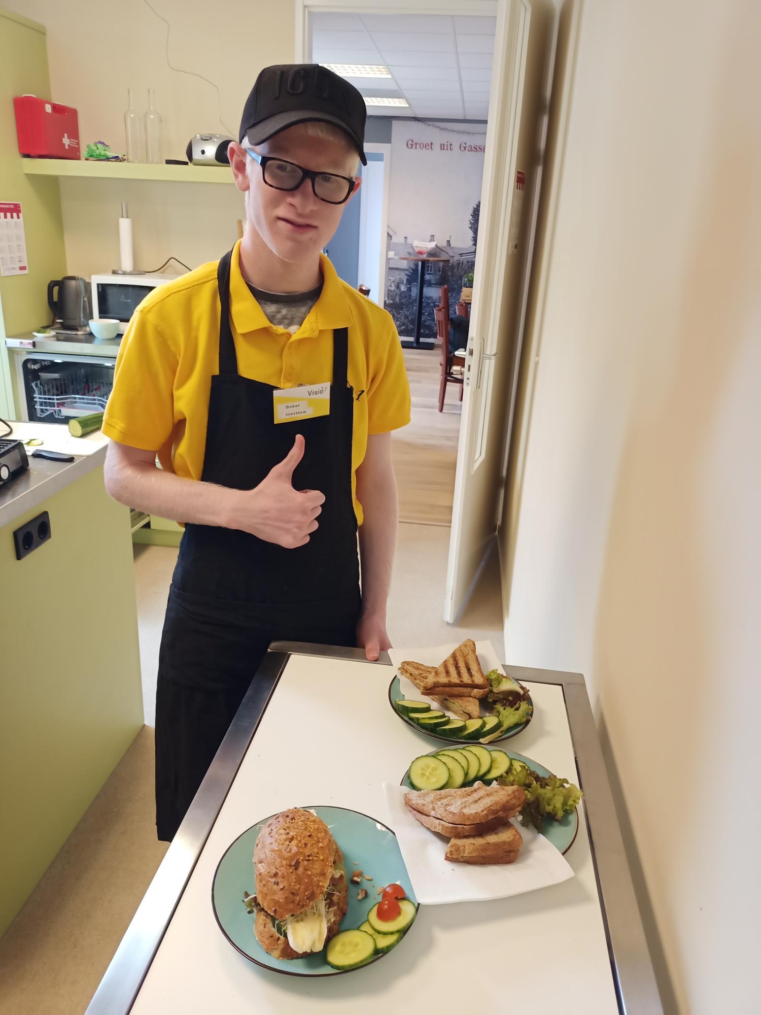 Leerling die bij Visit werkt, poseert lachend bij borden met belegde broodjes en steekt zijn duim op