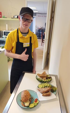 Leerling die bij Visit werkt, poseert lachend bij borden met belegde broodjes en steekt zijn duim op Leerling die bij Visit werkt, poseert lachend bij borden met belegde broodjes en steekt zijn duim op