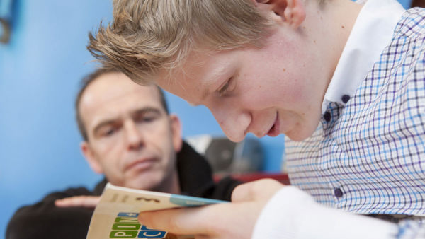 Leerling kijkt in een boek terwijl een docent meekijkt. Leerling kijkt in een boek terwijl een docent meekijkt.