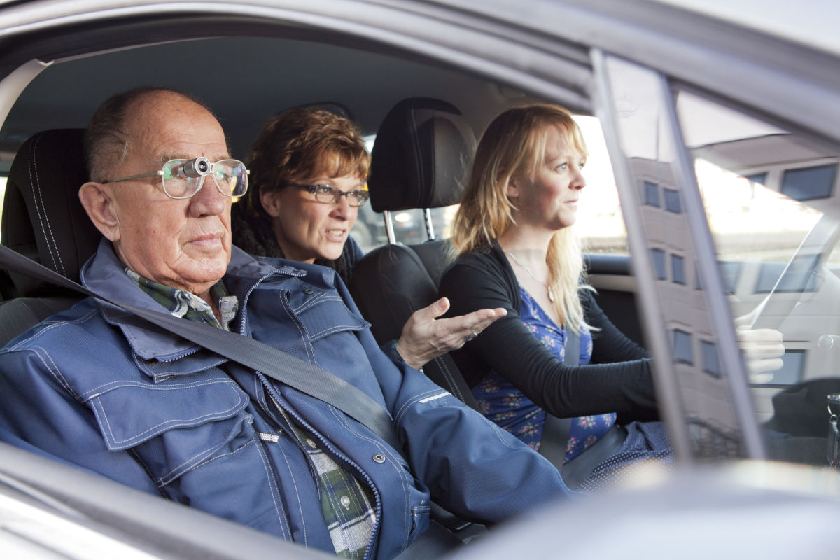 Drie personen zitten in een auto, de bijrijder draagt een bioptisch telescoopsysteem