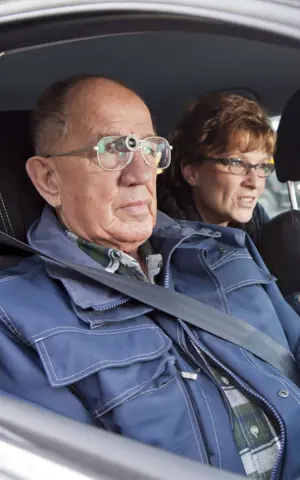 Drie personen zitten in een auto, de bijrijder draagt een bioptisch telescoopsysteem Drie personen zitten in een auto, de bijrijder draagt een bioptisch telescoopsysteem