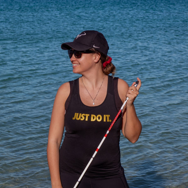 Marie-Elise poseert met haar taststok in een sportieve outfit met de tekst "Just do it" voor een meer met bergen op de achtergrond Marie-Elise poseert met haar taststok in een sportieve outfit met de tekst "Just do it" voor een meer met bergen op de achtergrond