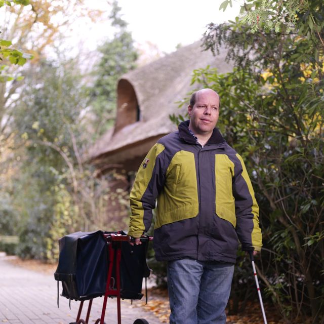 Cliënt brengt de post rond met een taststok in zijn hand en aan zijn andere hand een postwagentje Cliënt brengt de post rond met een taststok in zijn hand en aan zijn andere hand een postwagentje
