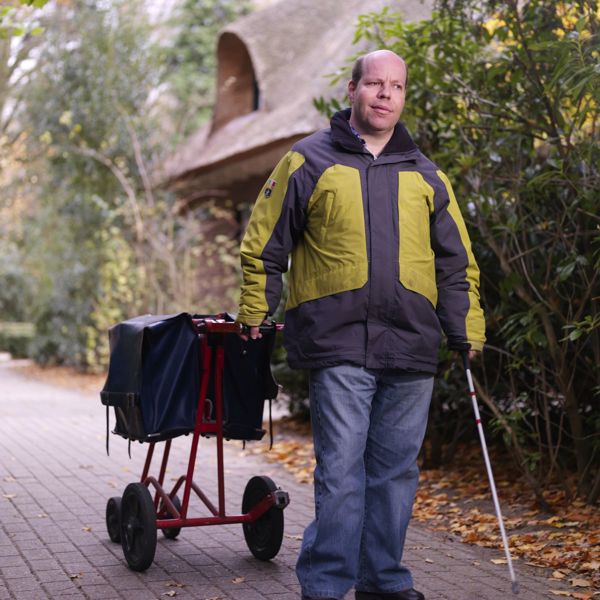 Cliënt brengt de post rond met een taststok in zijn hand en aan zijn andere hand een postwagentje Cliënt brengt de post rond met een taststok in zijn hand en aan zijn andere hand een postwagentje