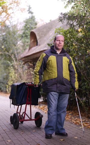 Cliënt brengt de post rond met een taststok in zijn hand en aan zijn andere hand een postwagentje Cliënt brengt de post rond met een taststok in zijn hand en aan zijn andere hand een postwagentje