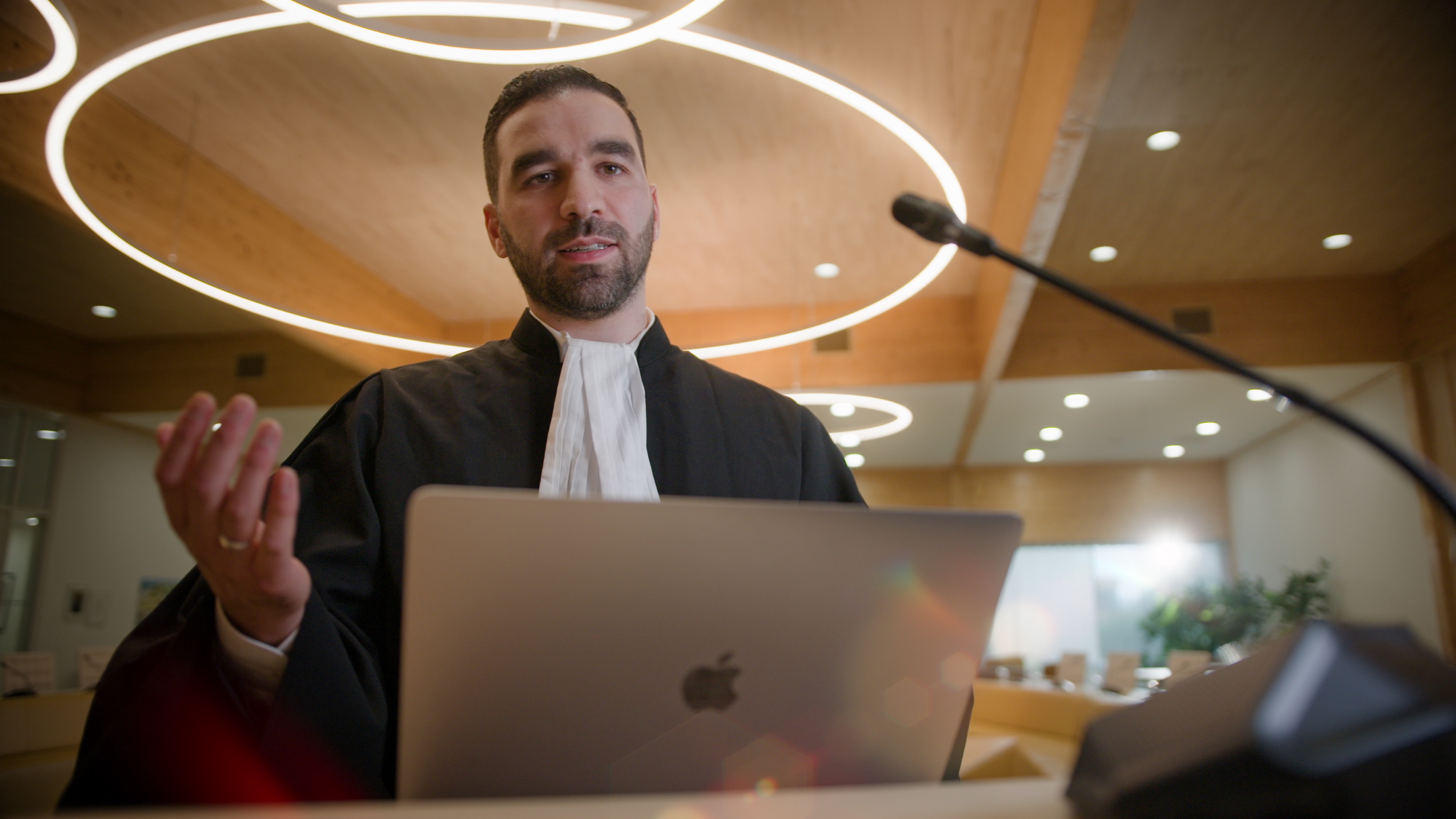 Faruk draagt zijn werkkleding (advocaat) en staat achter een microfoon. Zijn laptop staat voor hem. Hij is aan het praten. 