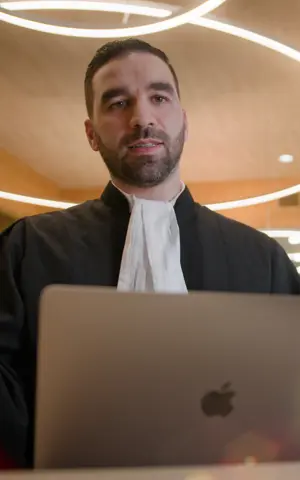 Faruk draagt zijn werkkleding (advocaat) en staat achter een microfoon. Zijn laptop staat voor hem. Hij is aan het praten. Faruk draagt zijn werkkleding (advocaat) en staat achter een microfoon. Zijn laptop staat voor hem. Hij is aan het praten.